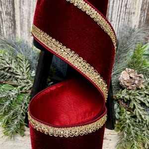 May include: A roll of deep red velvet ribbon featuring a metallic gold decorative trim along the edges. The ribbon is wound in a spiral shape, showcasing the soft texture of the fabric and the intricate, repeating pattern of the gold border.
