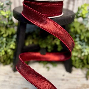 May include: A spool of red satin ribbon with a shimmery finish. The ribbon is wound around a black spool and is displayed on a wooden stool.