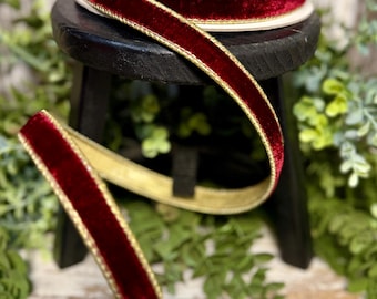 May include: A spool of burgundy velvet ribbon with a gold trim sits atop a small black stool. A length of the ribbon is unraveled, displaying its rich color and texture. The ribbon is a decorative element, suitable for crafts and embellishments.