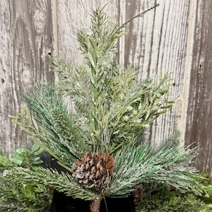 May include: A decorative artificial floral spray featuring a mix of green pine needles, slender evergreen branches, and small round berries. The foliage is lightly dusted with white artificial snow and features a single brown pinecone at the base.