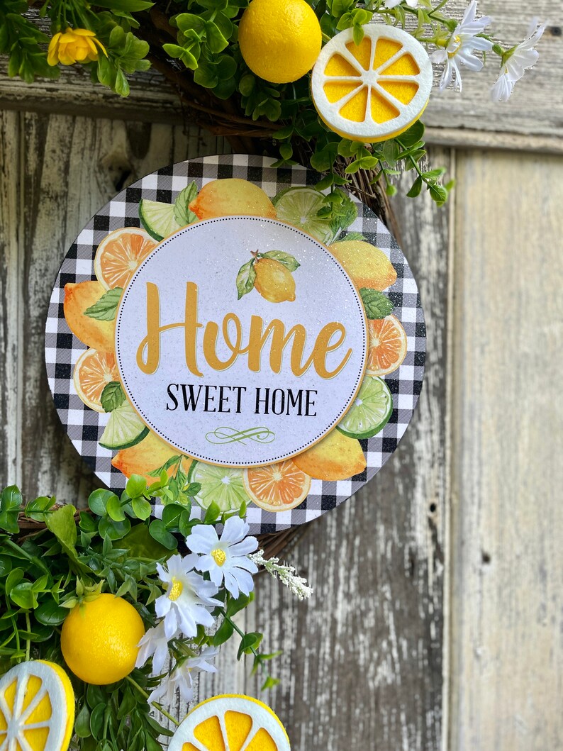 Może przedstawiać: Wieniec w czarno-białe kratę z okrągłym znakiem, na kt&oacute;rym napisano "Home Sweet Home", otoczony cytrynami i limonkami. Wieniec jest ozdobiony białymi kwiatami i zielonymi liśćmi.