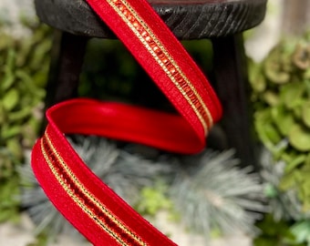 May include: A spool of red ribbon with gold trim and a central line of small, red embellishments. The ribbon is displayed on a small, black wooden stool, with greenery in the background. This decorative ribbon is suitable for crafts and gift wrapping.
