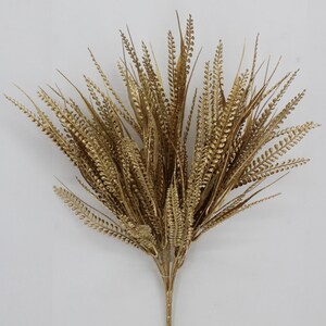May include: A decorative artificial plant spray featuring numerous slender, metallic gold stems with textured, fern-like foliage. The arrangement fans outward from a single central base, showcasing a uniform, shimmering gold finish across all leaves.