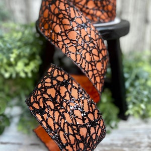 May include: Orange and black ribbon with a black spiderweb design and glitter. The ribbon is curled and displayed on a wooden surface.