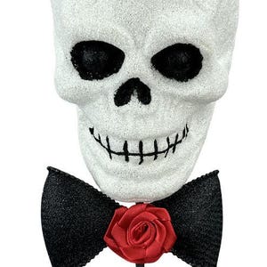 May include: A white glittery skull decoration on a thin black stick. The skull wears a black top hat adorned with a red fabric rose and a black glittery bow tie featuring a matching red fabric rose at the center. The skull has black eye sockets.
