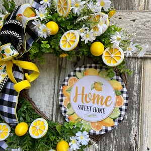 Może przedstawiać: Wianek w kształcie cytryny z czarno-białą wstążką w kratkę, białymi stokrotkami i okrągłym znakiem z napisem "Home Sweet Home". Znak jest otoczony cytrynami, limonkami i pomarańczami.
