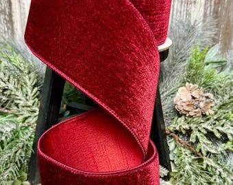 May include: A wide spool of deep red velvet ribbon with a soft, textured pile and finished edges. The ribbon is wound around a central core, with a portion of the fabric cascading downward in a loose, elegant spiral loop, showcasing the rich color.