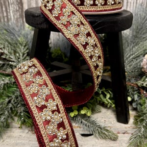 May include: A spool of deep red velvet ribbon featuring a decorative gold metallic border and a central pattern of small, clear crystal-like floral embellishments with white pearl centers, arranged in a repeating sequence along the length.