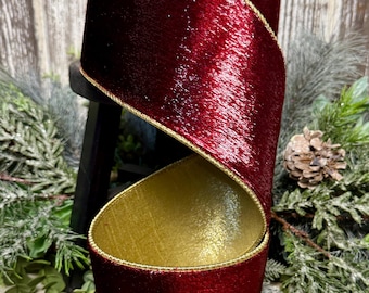 May include: A wide spool of deep red velvet ribbon featuring a metallic gold underside and gold-trimmed edges. The ribbon is coiled in a spiral shape, showcasing the contrast between the dark red textured velvet exterior and the shimmering gold interior.