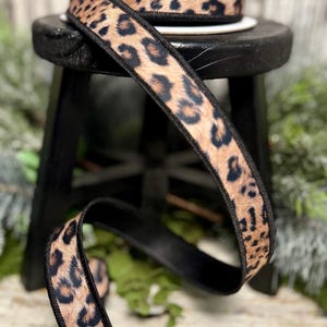 May include: A roll of leopard print ribbon with a black border sits on a small black stool. A length of the ribbon is unrolled, showing the tan and black animal print. The ribbon is likely used for crafting or gift wrapping.