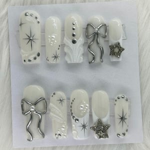 May include: A set of ten artificial nails in various designs. The nails are primarily white with silver accents, including bows, stars, and decorative embellishments. Some nails feature starburst designs and pearl-like details.