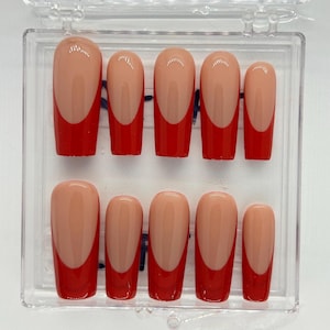 Pode incluir: Um conjunto de dez unhas artificiais com um design de manicure francesa. As unhas têm uma base cor de pele com uma ponta vermelha brilhante. As unhas são exibidas em uma caixa de plástico transparente.