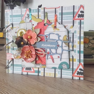 May include: A handmade scrapbook with a railway theme. The cover features a patterned paper with train crossing signs and a tag with the text "#here&now". Embellishments include paper flowers in shades of peach and brown.