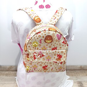 May include: A small beige backpack with a front pocket. The backpack features a woodland animal print with foxes, owls, and mushrooms in shades of red, orange, brown, and green.