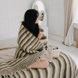 May include: A woman sits on a bed wrapped in a striped blanket, holding a book. The blanket is a mix of brown and white stripes with a fringe edge. The bed is covered in a white and beige blanket.