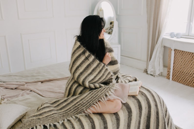 May include: A woman sits on a bed wrapped in a striped blanket, holding a book. The blanket is a mix of brown and white stripes with a fringe edge. The bed is covered in a white and beige blanket.