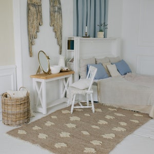May include: A white bedroom with a bed, a white chair, a wooden table, a mirror, a wicker basket, and a rug with a cloud pattern. The walls are white and there are wooden angel wings on the wall. The bed has a white headboard and a white blanket. The chair is white and has a white cushion. The table is wooden and has a white top. The mirror is round and has a gold frame. The basket is wicker and is empty. The rug is brown and has a white cloud pattern.