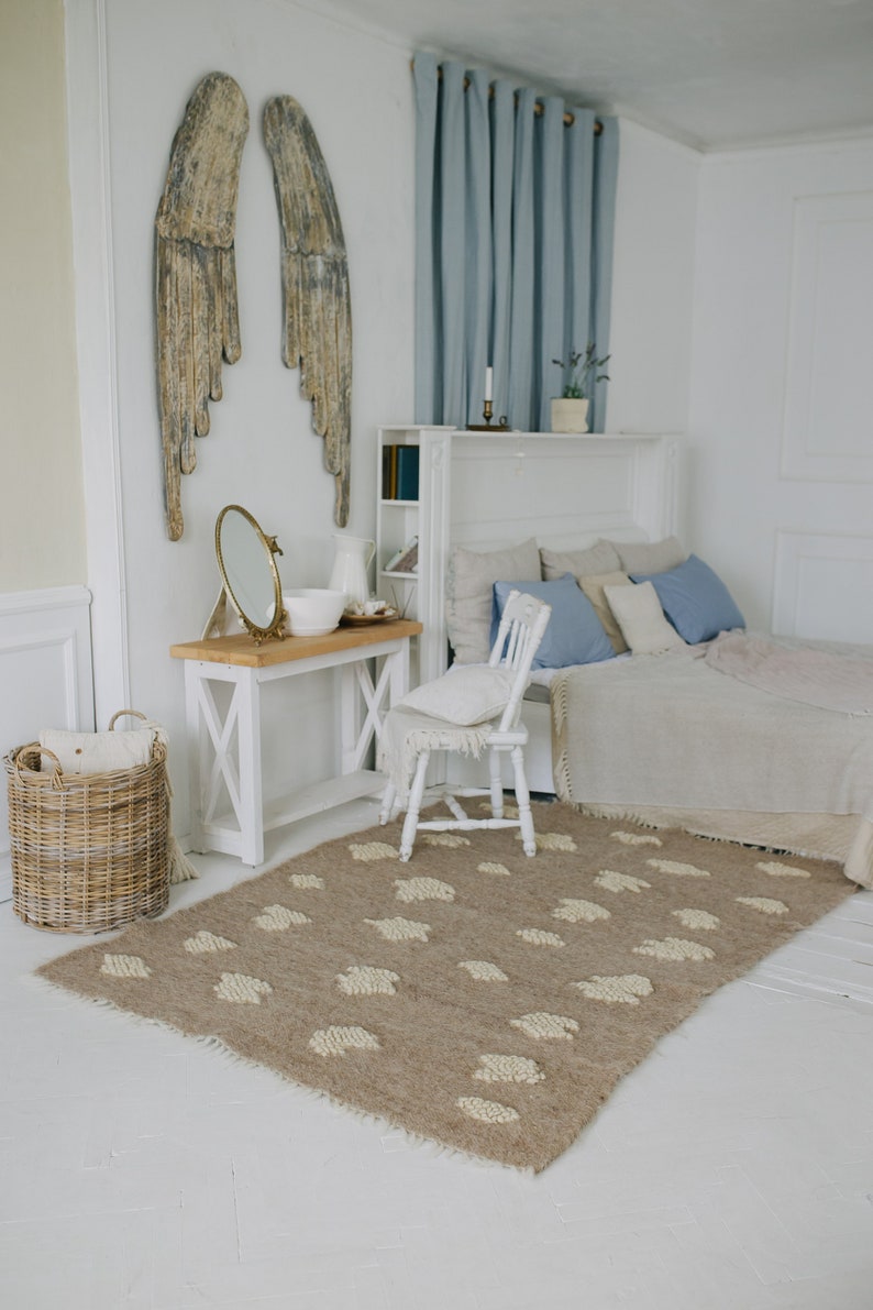 May include: A white bedroom with a bed, a white chair, a wooden table, a mirror, a wicker basket, and a rug with a cloud pattern. The walls are white and there are wooden angel wings on the wall. The bed has a white headboard and a white blanket. The chair is white and has a white cushion. The table is wooden and has a white top. The mirror is round and has a gold frame. The basket is wicker and is empty. The rug is brown and has a white cloud pattern.