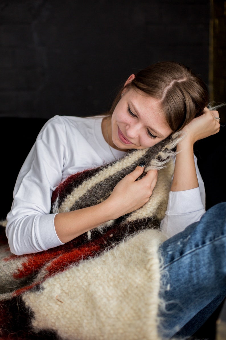 May include: A young woman is sitting with a white long-sleeved shirt and blue jeans. She is holding a brown, white, and red striped blanket.