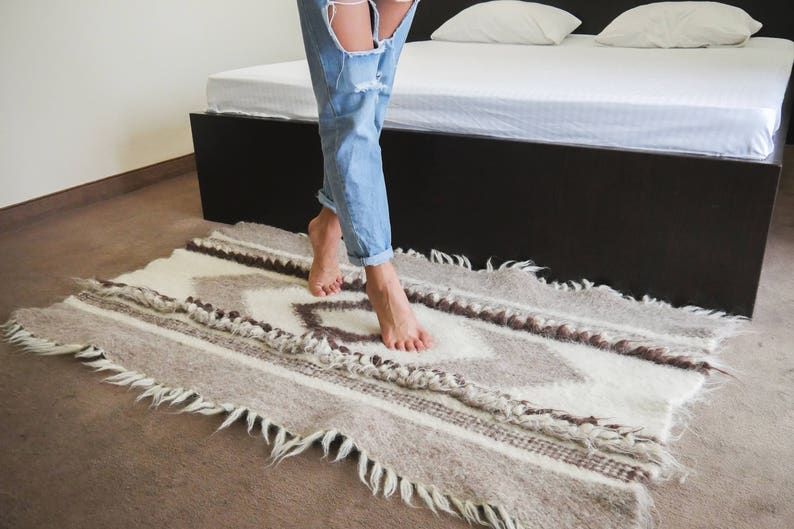 May include: A person's feet are standing on a white and brown patterned rug with fringe. The rug is on a wooden floor. The person is wearing blue jeans.