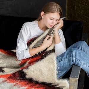 May include: A young person is sitting on a couch, wrapped in a colorful blanket with red, white, and brown stripes. They are resting their head on the blanket and have their eyes closed.