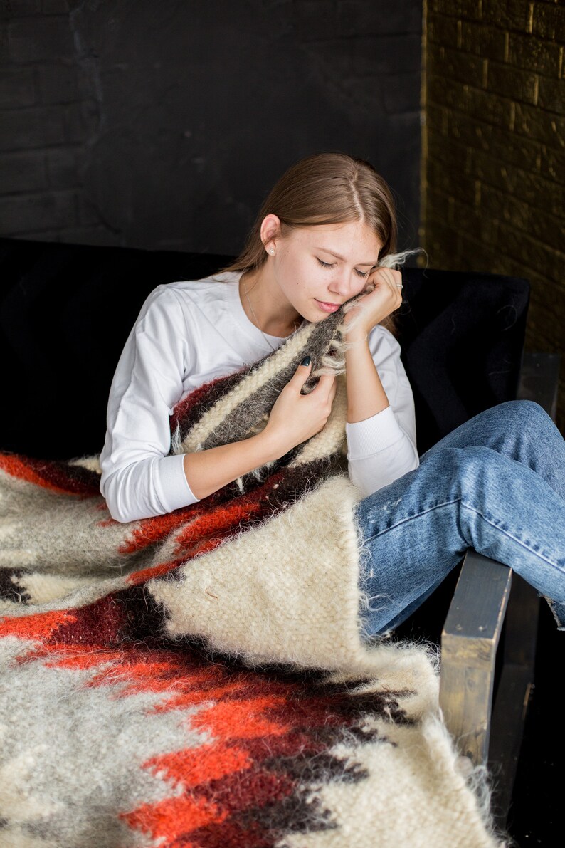 May include: A young person is sitting on a couch, wrapped in a colorful blanket with red, white, and brown stripes. They are resting their head on the blanket and have their eyes closed.