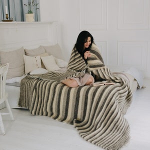 May include: A woman sits on a bed with a striped blanket, reading a book. The bed has a white headboard and is made with white linens. The room is decorated in a minimalist style with white walls and a white chair.