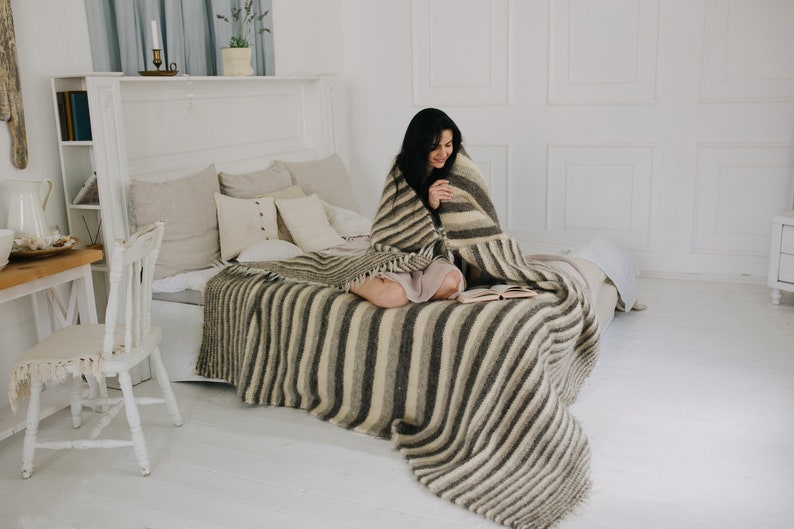 May include: A woman sits on a bed with a striped blanket, reading a book. The bed has a white headboard and is made with white linens. The room is decorated in a minimalist style with white walls and a white chair.