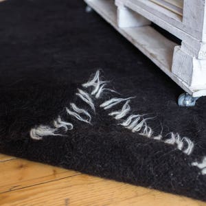 May include: A black fuzzy rug with white fringe edges is partially covering a white wooden pallet with wheels. The pallet is on a wooden floor.