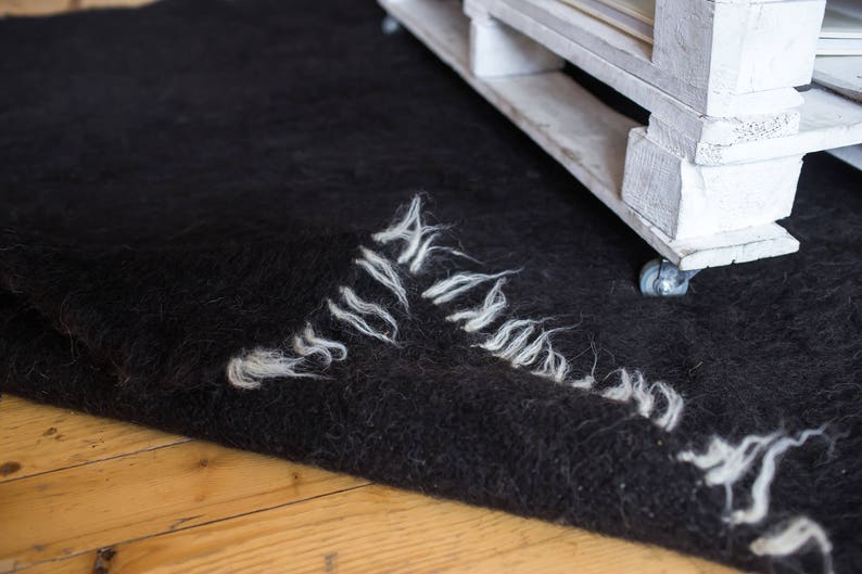 May include: A black fuzzy rug with white fringe edges is partially covering a white wooden pallet with wheels. The pallet is on a wooden floor.