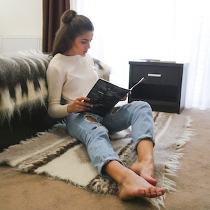 May include: A woman sitting on a bed with a fuzzy gray and white blanket, reading a magazine. She is wearing a white sweater and blue jeans.
