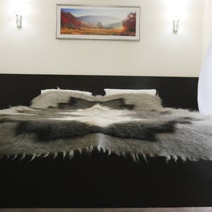May include: A plush, gray and white blanket with a fringed edge, folded on a black bed frame. The bed is in a bedroom with white walls and a window with white curtains.