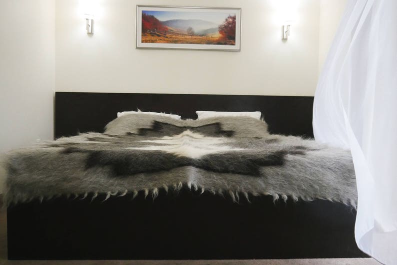 May include: A plush, gray and white blanket with a fringed edge, folded on a black bed frame. The bed is in a bedroom with white walls and a window with white curtains.