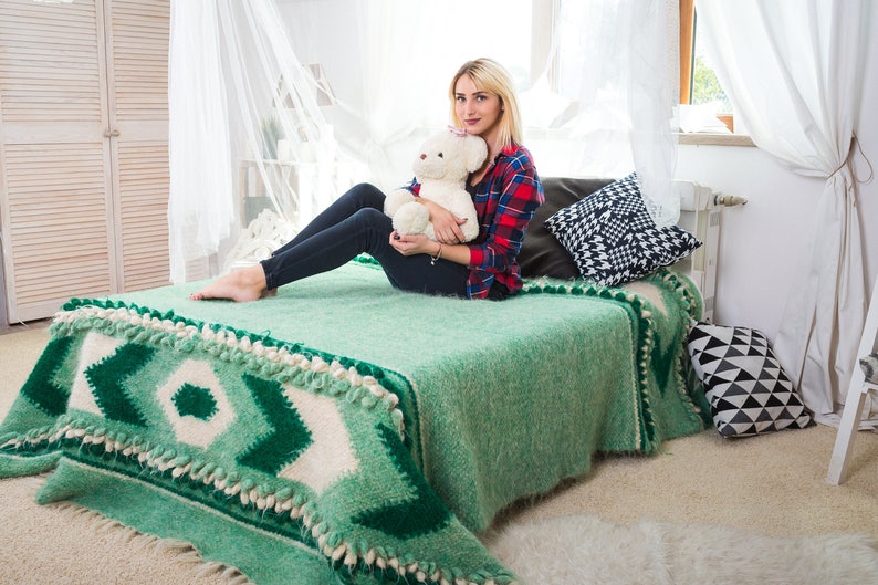 May include: A woman sits on a bed with a green and white patterned blanket. She is holding a white teddy bear and is wearing a red and black plaid shirt and blue jeans.