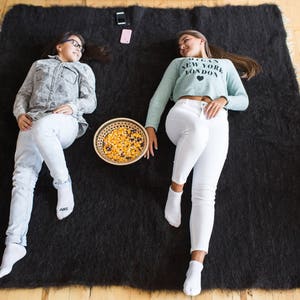 May include: Two people are relaxing on a black fuzzy rug. One person is wearing a blue and white patterned shirt and white jeans. The other person is wearing a light blue shirt with the text "I Love New York London" and white jeans. A bowl of snacks is in the center of the rug.