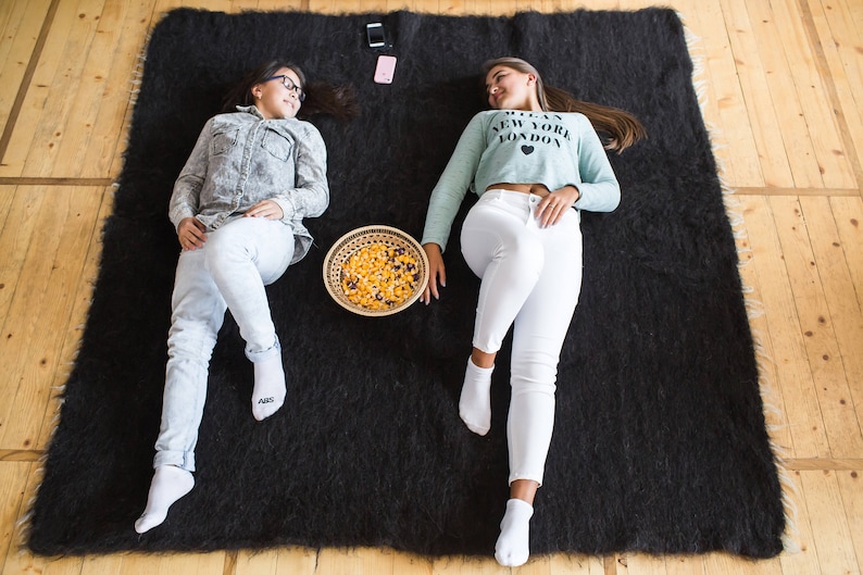 May include: Two people are relaxing on a black fuzzy rug. One person is wearing a blue and white patterned shirt and white jeans. The other person is wearing a light blue shirt with the text "I Love New York London" and white jeans. A bowl of snacks is in the center of the rug.