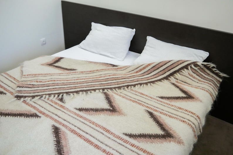 May include: A white bed with a brown headboard and two white pillows. A white and brown patterned blanket is draped over the bed.