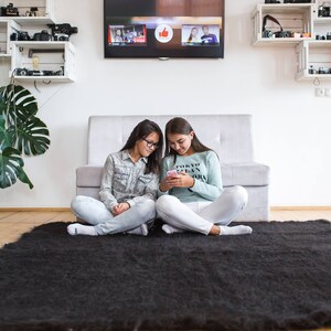 May include: Two people are sitting on a black shag rug in front of a white couch. They are looking at a phone and smiling. The couch is in front of a white wall with shelves on either side. The shelves are filled with cameras and other items.