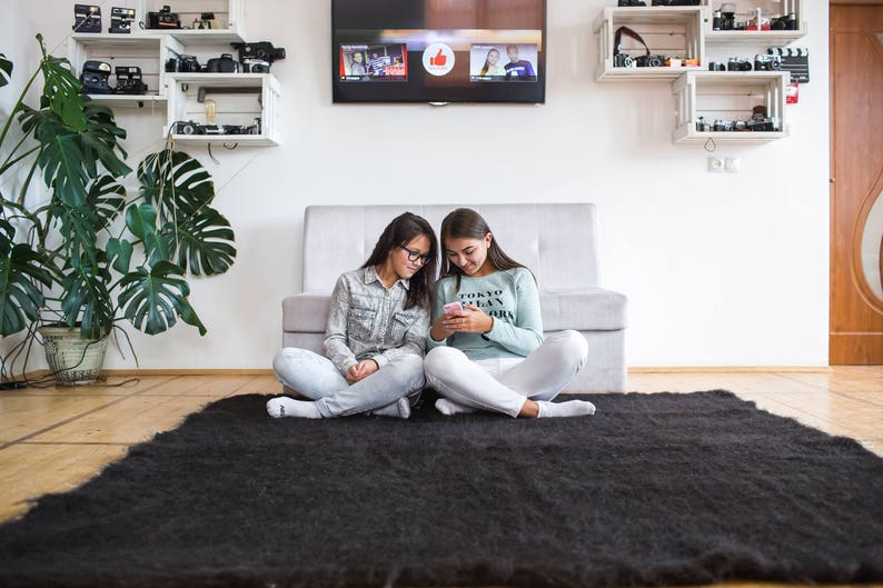 May include: Two people are sitting on a black shag rug in front of a white couch. They are looking at a phone and smiling. The couch is in front of a white wall with shelves on either side. The shelves are filled with cameras and other items.