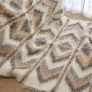 Puede incluir: Una manta blanca, marrón y gris peluda con un patrón geométrico de chevron. La manta está hecha de fibras suaves y esponjosas y tiene un borde con flecos.