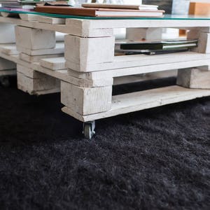 May include: White pallet coffee table with a glass top and wheels. The table has three shelves and is sitting on a black rug.