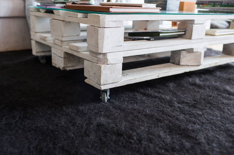 May include: White pallet coffee table with a glass top and wheels. The table has three shelves and is sitting on a black rug.