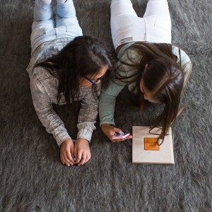 May include: Two people are lying on a gray rug, looking at a small phone. One person is wearing white pants and a white shirt, the other is wearing blue jeans and a gray shirt.