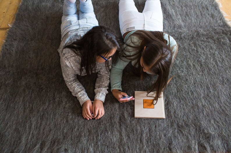 May include: Two people are lying on a gray rug, looking at a small phone. One person is wearing white pants and a white shirt, the other is wearing blue jeans and a gray shirt.