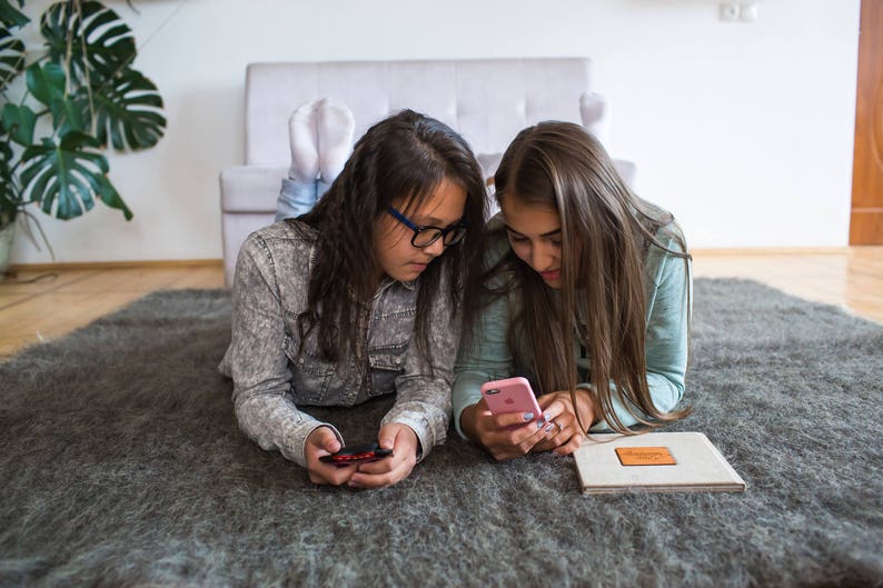 May include: Two young people are lying on a gray rug, looking at their phones. One person is wearing glasses and a gray shirt, while the other person is wearing a blue shirt. They are both smiling.
