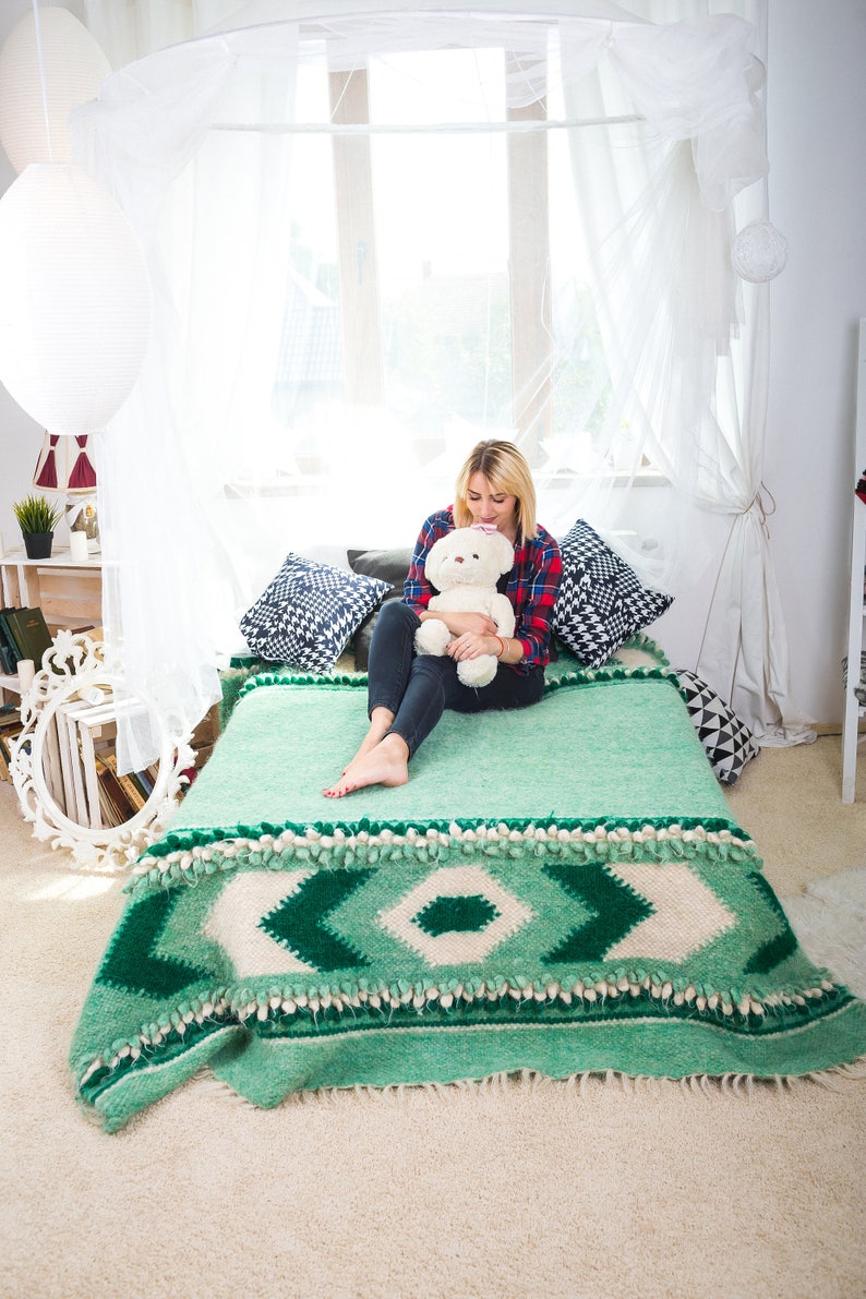 May include: A woman sits on a bed with a green and white patterned blanket. She is holding a white teddy bear. The bed has a white canopy and there are pillows on the bed.