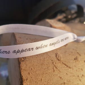 May include: A white ribbon with the text "feathers appear when angels are near" printed in dark grey. The ribbon is knotted and lies on a wooden surface.