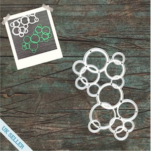 May include: Metal die cut in the shape of a cluster of overlapping circles. The die cut is silver and has a smooth, shiny finish. The die cut is on a wooden background.
