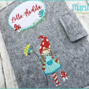Puede incluir: Una cubierta de libro de fieltro gris con un nombre bordado personalizado "Lotta-Madita" en un óvalo blanco. La cubierta también presenta una ilustración bordada de una niña con cabello rojo, un sombrero de lunares rojos y blancos y un vestido azul que sostiene una flor amarilla. El texto "mariLena Stoffzauberer" está bordado en la esquina superior derecha.