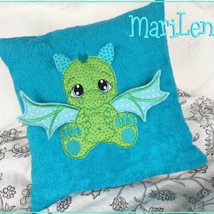 Könnte beinhalten: Ein türkisblaues Kissen mit einem grünen Drachen, der darauf gestickt ist. Der Drache hat große Augen und einen hellblauen Flügel. Das Kissen hat den Text "mariLena" aufgestickt.